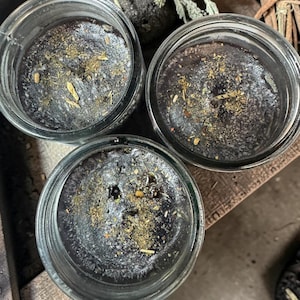 May include: Three glass jars containing black wax candles. The surface of each candle is dark black and topped with a scattering of dried herbs and botanical particles. Each candle features a single centered wick visible within the clear glass.