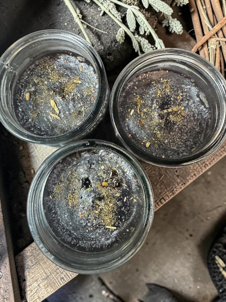 May include: Three glass jars containing black wax candles. The surface of each candle is dark black and topped with a scattering of dried herbs and botanical particles. Each candle features a single centered wick visible within the clear glass.