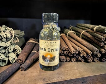 May include: A clear glass bottle of 4 Lunas Botanica Road Opener ritual oil, 1 oz size, featuring a label with a sunrise graphic over a road. The bottle contains liquid and herbs, sealed with a plastic cap, positioned between bundles of cinnamon sticks.
