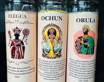 May include: Three glass jar candles with labels featuring the names Elegua, Ochun, and Orula. Each candle has a different color at the bottom and includes text and illustrations. The candles are from 4 Lunas Botanica.