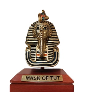 Puede incluir: Una máscara decorativa dorada de Tutankamón, con detalles en negro y azul, montada sobre una base de madera. La base tiene una placa dorada que dice "MASK OF TUT". La máscara presenta una elaborada tocado.
