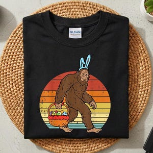May include: Black t-shirt featuring a Bigfoot character wearing bunny ears and carrying a basket of Easter eggs. The Bigfoot is brown, set against a retro sunset design. The shirt is a Gildan brand.