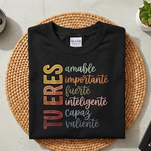 Puede incluir: Camiseta negra de cuello redondo con las palabras "TU ERES" en texto vertical y colorido. Debajo, las palabras "amable, importante, fuerte, inteligente, capaz, valiente" están escritas en una combinación de colores a juego. La camiseta está sobre una esterilla tejida.