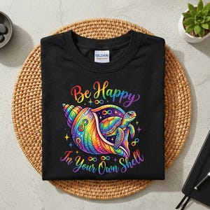 Könnte beinhalten: Schwarzes T-Shirt mit einem farbenfrohen Motiv einer Meeresschildkröte in einer Regenbogen-Muschel. Der Text "Be Happy In Your Own Shell" steht oberhalb und unterhalb des Motivs in Regenbogenfarben.