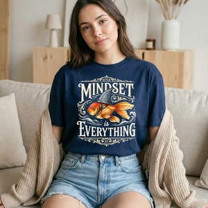 Peut inclure: T-shirt bleu marine avec l'inscription blanche « MINDSET is EVERYTHING ». Le motif représente un poisson rouge portant un aileron de requin. Le t-shirt est porté avec un short en jean et un cardigan beige.