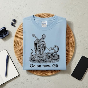 Op de afbeelding: Lichtblauw T-shirt met een zwart-wit illustratie van een geklede figuur met een staf, die vecht tegen twee slangen. De tekst "Go on now. Git." staat eronder. Het shirt is gemaakt van zacht materiaal.