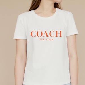 Puede incluir: Camiseta blanca con la palabra "COACH" en letras naranjas sobre "NEW YORK". La camiseta tiene cuello redondo y mangas cortas. La tela parece suave.