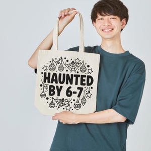 May include: A cream-colored tote bag with the words "HAUNTED BY 6-7" in black, surrounded by Halloween-themed illustrations. The bag is held by a person wearing a teal t-shirt.