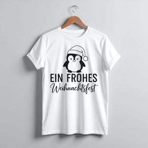 May include: White t-shirt featuring a black and white penguin wearing a Santa hat. The text "EIN FROHES Weihnachtsfest" is printed below the penguin. The shirt is hanging on a wooden hanger.