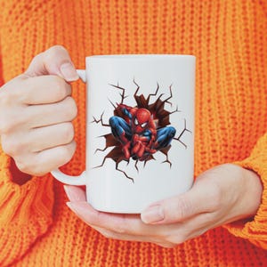 Peut inclure: Mug en c&eacute;ramique blanche avec un motif de Spider-Man en rouge et bleu, sortant d'un mur fissur&eacute;. La tasse a une anse incurv&eacute;e. La personne porte un pull orange.