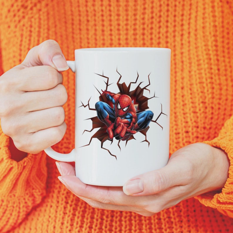 Peut inclure: Mug en c&eacute;ramique blanche avec un motif de Spider-Man en rouge et bleu, sortant d'un mur fissur&eacute;. La tasse a une anse incurv&eacute;e. La personne porte un pull orange.
