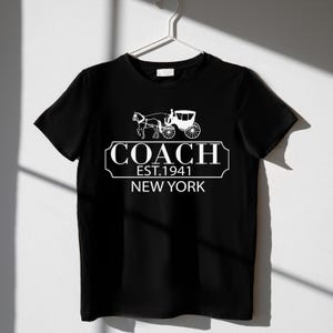 Puede incluir: Camiseta negra con un gr&aacute;fico blanco de un carruaje tirado por caballos y el texto "COACH EST.1941 NEW YORK". La camiseta est&aacute; colgada en una percha blanca sobre un fondo blanco.