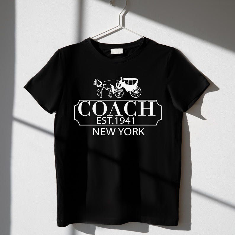 Puede incluir: Camiseta negra con un gr&aacute;fico blanco de un carruaje tirado por caballos y el texto "COACH EST.1941 NEW YORK". La camiseta est&aacute; colgada en una percha blanca sobre un fondo blanco.