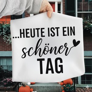 Peut inclure: Sac fourre-tout en toile blanche avec l'expression allemande "...HEUTE IST EIN sch&ouml;ner TAG" imprim&eacute;e en noir. Un c&oelig;ur noir est &agrave; c&ocirc;t&eacute; du mot "sch&ouml;ner". Le sac est tenu devant un b&acirc;timent en briques.