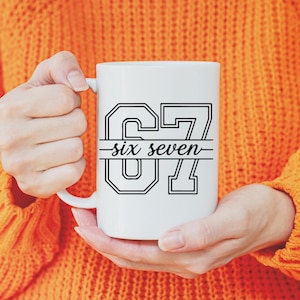May include: White ceramic mug with the text "67 six seven" in a black outlined font. The mug is held by a person wearing an orange sweater. The mug is a standard size for coffee or tea.
