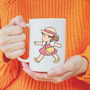 May include: White ceramic mug featuring a cartoon illustration of a girl in a pink dress, yellow purse, and straw hat. The girl is walking with a determined expression. The mug has a white handle and is held by a person wearing an orange sweater.