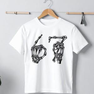 May include: White t-shirt with a black graphic of two skeleton hands forming the numbers 6 and 7. The shirt is hanging on a wooden hanger against a light gray background.