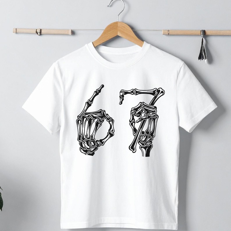 May include: White t-shirt with a black graphic of two skeleton hands forming the numbers 6 and 7. The shirt is hanging on a wooden hanger against a light gray background.