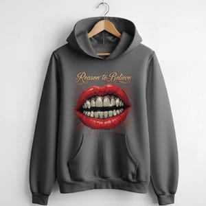May include: A charcoal gray hooded sweatshirt featuring a graphic print of bright red lips with diamond-encrusted teeth. Above the lips, the text Reason to Believe is printed in a gold cursive font. The hoodie includes a front kangaroo pocket.