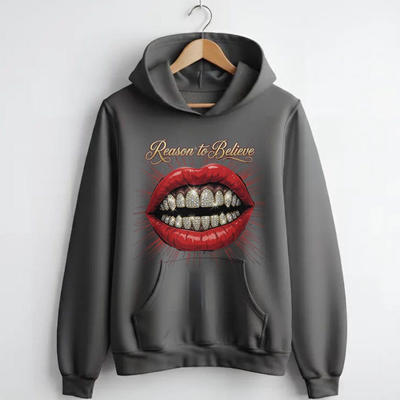 May include: A charcoal gray hooded sweatshirt featuring a graphic print of bright red lips with diamond-encrusted teeth. Above the lips, the text Reason to Believe is printed in a gold cursive font. The hoodie includes a front kangaroo pocket.