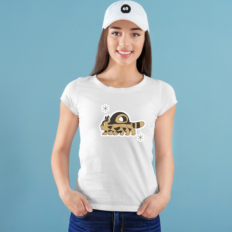 May include: White t-shirt featuring a cartoon caterpillar design in shades of brown and tan, with a dark brown shell. The design is accompanied by two white snowflake accents. The model is wearing a white baseball cap.