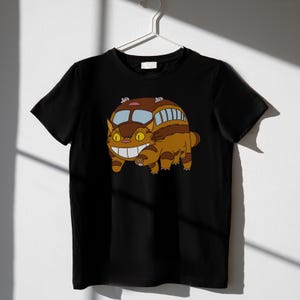 May include: Black t-shirt featuring a cartoon illustration of a cat-bus character. The cat-bus is brown and tan with a smiling face and bus-like features. The design is centered on the shirt.