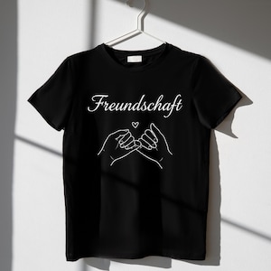 Peut inclure: T-shirt noir avec le mot "Freundschaft" en &eacute;criture cursive et un dessin au trait blanc de deux mains se faisant une promesse de petit doigt, avec un petit c&oelig;ur au-dessus. Le t-shirt est suspendu &agrave; un cintre blanc.