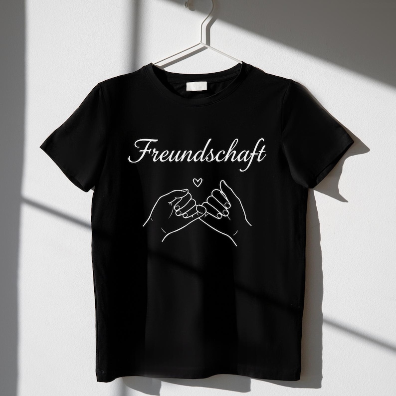 Peut inclure: T-shirt noir avec le mot "Freundschaft" en &eacute;criture cursive et un dessin au trait blanc de deux mains se faisant une promesse de petit doigt, avec un petit c&oelig;ur au-dessus. Le t-shirt est suspendu &agrave; un cintre blanc.