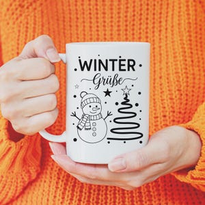 May include: White ceramic mug featuring a snowman design with a hat, scarf, and a Christmas tree. The mug has the words "Winter Gr&uuml;&szlig;e" in black script. The mug is held by a person wearing an orange sweater.