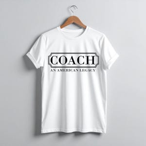 Puede incluir: Camiseta blanca con la palabra "COACH" en un marco rectangular negro. Debajo, el texto "AN AMERICAN LEGACY" en negro. La camiseta est&aacute; colgada de una percha de madera.
