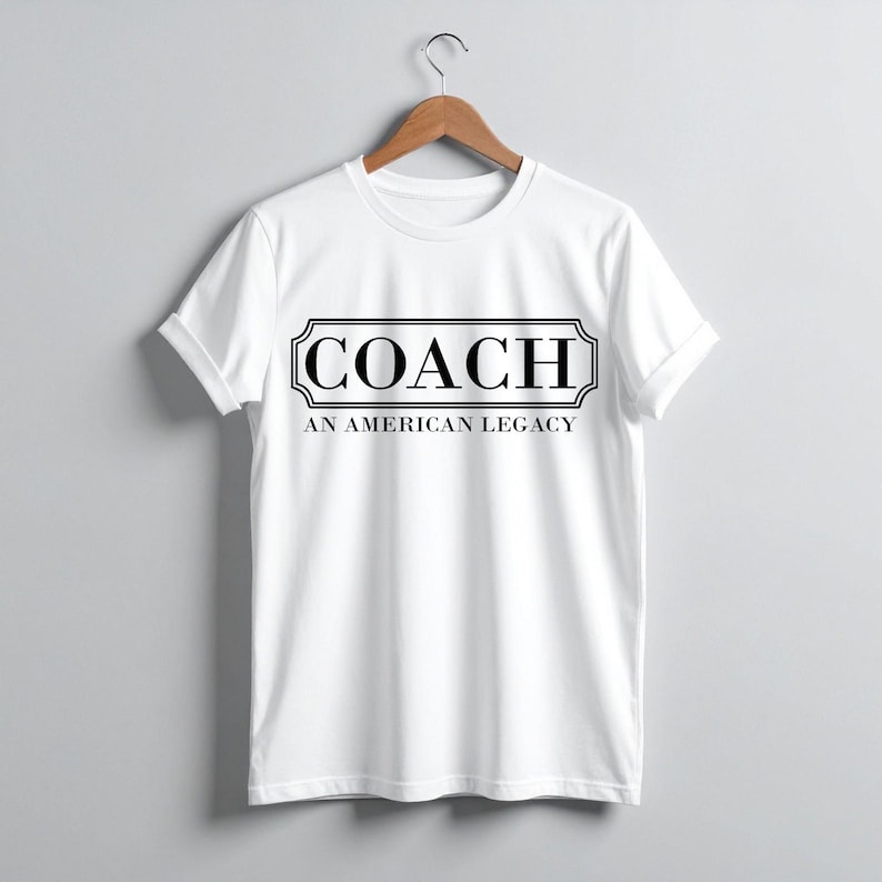Puede incluir: Camiseta blanca con la palabra "COACH" en un marco rectangular negro. Debajo, el texto "AN AMERICAN LEGACY" en negro. La camiseta est&aacute; colgada de una percha de madera.