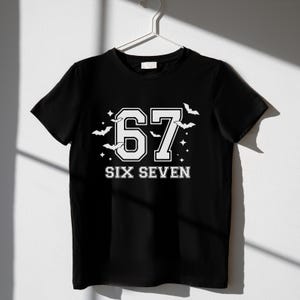 May include: Black t-shirt with the number "67" in white, outlined in black, and the words "SIX SEVEN" below. The design includes white bats and stars. The shirt is hanging on a white hanger.
