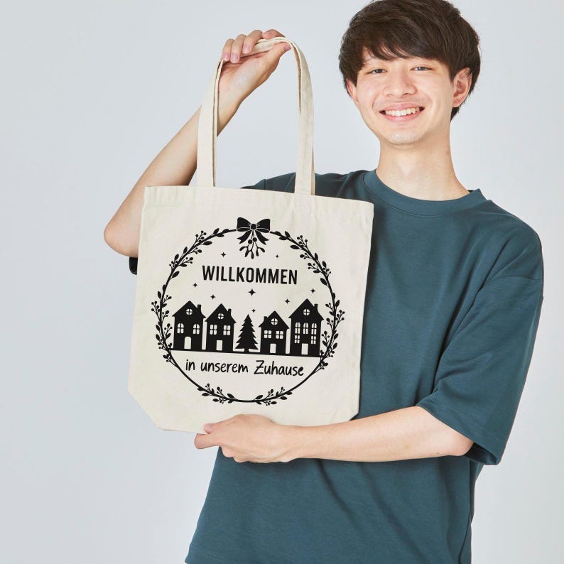 May include: A cream-colored tote bag with a black wreath design. The wreath surrounds the German word "Willkommen" and the phrase "in unserem Zuhause" with illustrations of houses and a Christmas tree.