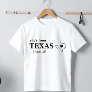 Pu&ograve; includere: T-shirt bianca con la scritta "She's from TEXAS I can tell" in nero. Un contorno nero dello stato del Texas con un cuore al centro &egrave; a destra del testo. La maglietta &egrave; appesa a una gruccia di legno.