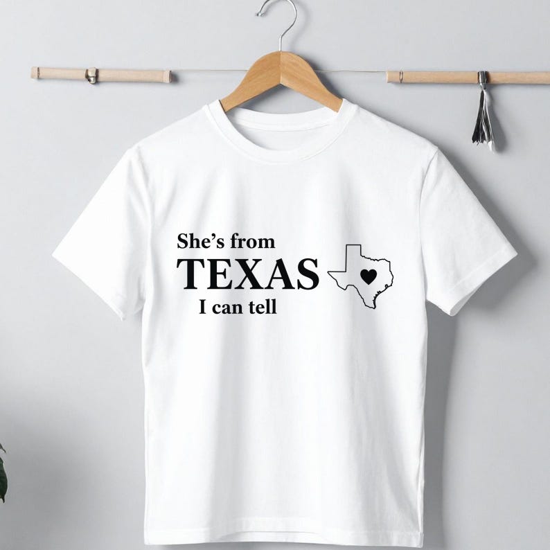 Pu&ograve; includere: T-shirt bianca con la scritta "She's from TEXAS I can tell" in nero. Un contorno nero dello stato del Texas con un cuore al centro &egrave; a destra del testo. La maglietta &egrave; appesa a una gruccia di legno.