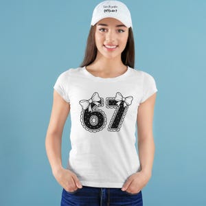 May include: White t-shirt with a black and white graphic of the numbers 6 and 7, each adorned with a bow. The shirt is paired with a white baseball cap that has the text "Solve the problem (97)+4=?" in black.