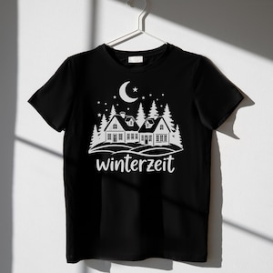 May include: Black t-shirt with a white graphic of a winter scene. The design includes houses, pine trees, a crescent moon, stars, and the word "winterzeit". The shirt is hanging on a white hanger.