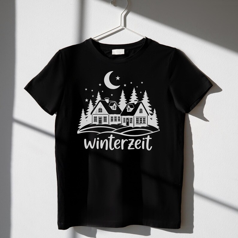May include: Black t-shirt with a white graphic of a winter scene. The design includes houses, pine trees, a crescent moon, stars, and the word "winterzeit". The shirt is hanging on a white hanger.