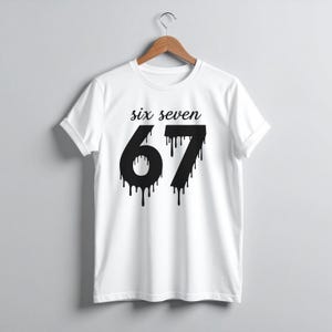 May include: White t-shirt with the words "six seven" above the number "67" in black, dripping font. The shirt is hanging on a wooden hanger against a light gray background. The sleeves are cuffed.