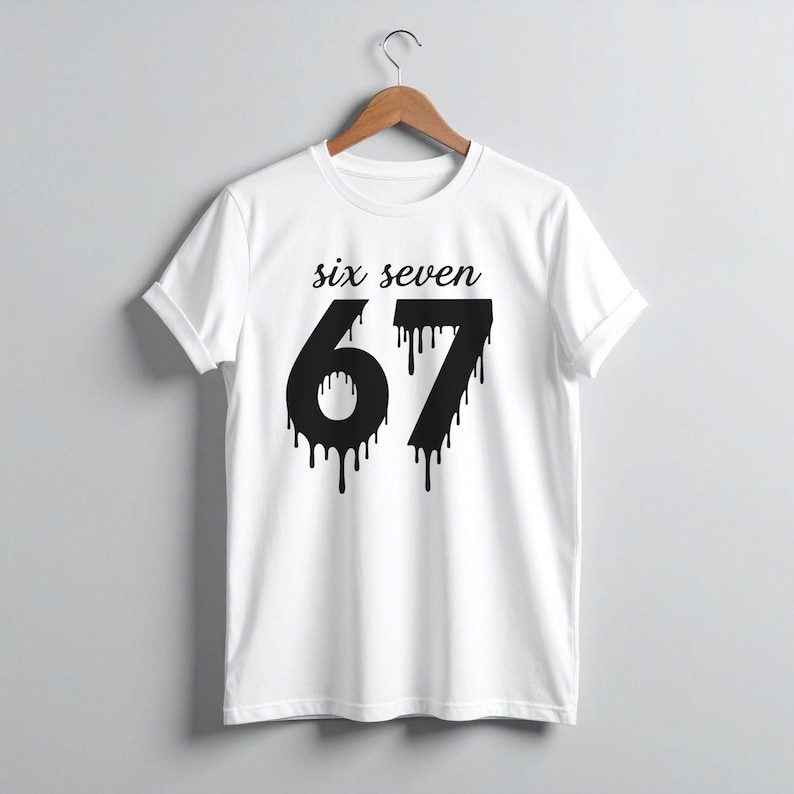 May include: White t-shirt with the words "six seven" above the number "67" in black, dripping font. The shirt is hanging on a wooden hanger against a light gray background. The sleeves are cuffed.