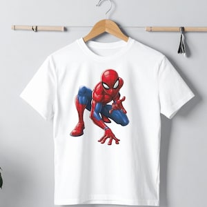 Peut inclure: T-shirt blanc avec une illustration color&eacute;e de Spider-Man en position accroupie. Le super-h&eacute;ros est repr&eacute;sent&eacute; dans son costume rouge et bleu classique. Le t-shirt est suspendu &agrave; un cintre en bois.