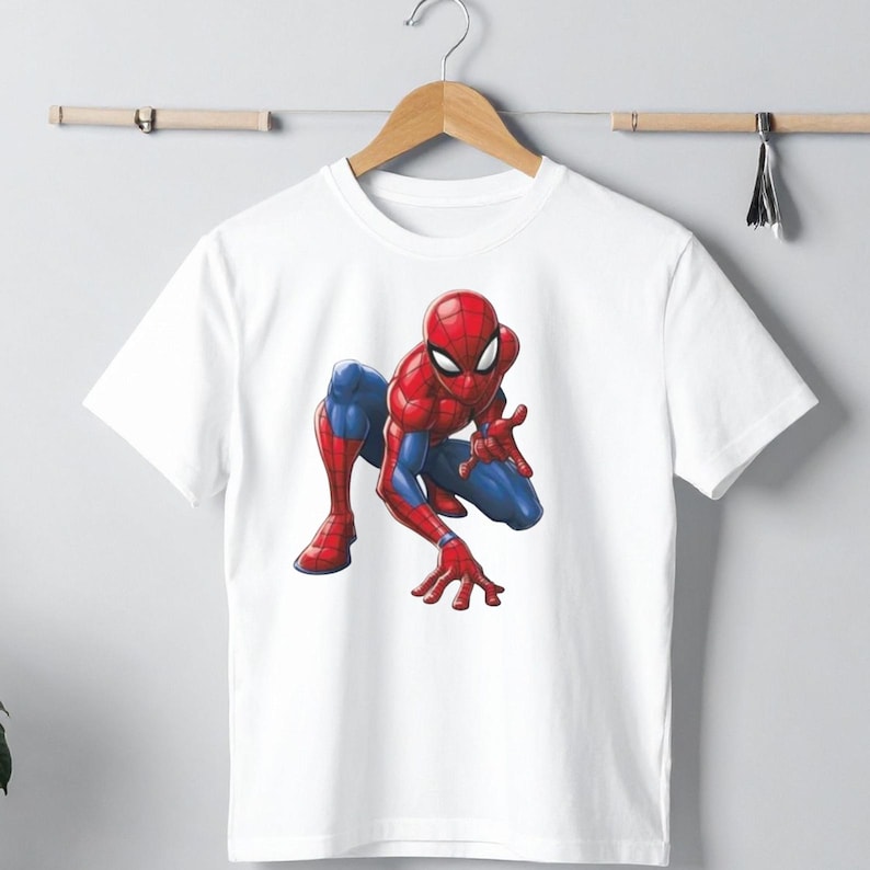 Peut inclure: T-shirt blanc avec une illustration color&eacute;e de Spider-Man en position accroupie. Le super-h&eacute;ros est repr&eacute;sent&eacute; dans son costume rouge et bleu classique. Le t-shirt est suspendu &agrave; un cintre en bois.