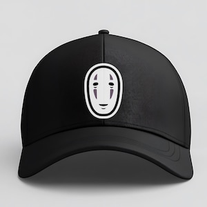 May include: Black baseball cap featuring a white and purple graphic of a character's face from the animated film, Spirited Away. The cap has a curved brim and a button on top.