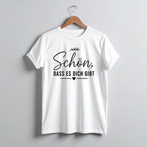 Peut inclure: T-shirt blanc avec l'inscription en allemand "Sch&ouml;n, dass es dich gibt" en noir, signifiant "Heureux que tu existes". Le design comprend un petit c&oelig;ur et des accents de feuilles. Le t-shirt est suspendu &agrave; un cintre en bois.
