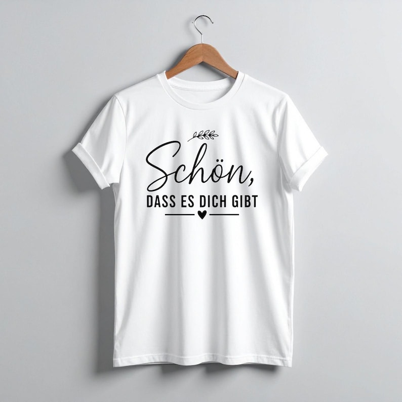 Peut inclure: T-shirt blanc avec l'inscription en allemand "Sch&ouml;n, dass es dich gibt" en noir, signifiant "Heureux que tu existes". Le design comprend un petit c&oelig;ur et des accents de feuilles. Le t-shirt est suspendu &agrave; un cintre en bois.