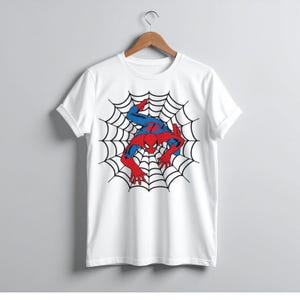 Peut inclure: T-shirt blanc avec un motif de Spider-Man dans une toile d'araign&eacute;e. Le super-h&eacute;ros est repr&eacute;sent&eacute; dans son costume rouge et bleu, avec une toile noire en arri&egrave;re-plan. Le t-shirt est suspendu &agrave; un cintre en bois.