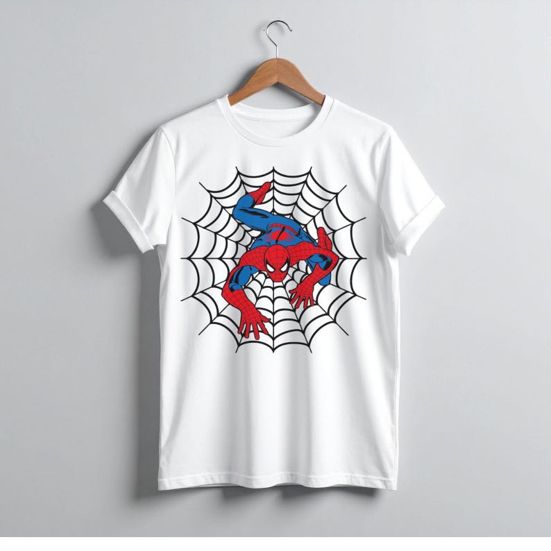 Peut inclure: T-shirt blanc avec un motif de Spider-Man dans une toile d'araign&eacute;e. Le super-h&eacute;ros est repr&eacute;sent&eacute; dans son costume rouge et bleu, avec une toile noire en arri&egrave;re-plan. Le t-shirt est suspendu &agrave; un cintre en bois.