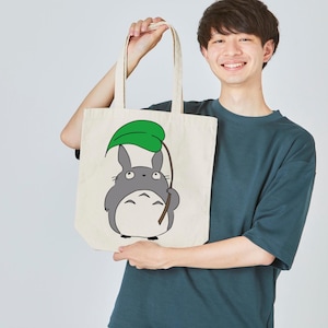 May include: A cream-colored tote bag featuring a cartoon illustration of Totoro holding a green leaf. The bag has long handles and is made of a sturdy fabric, perfect for carrying groceries or books.