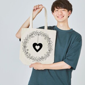 Peut inclure: Sac fourre-tout cr&egrave;me avec un pendentif en forme de c&oelig;ur noir &agrave; l'int&eacute;rieur d'une couronne florale. La couronne est compos&eacute;e de feuilles, de petites fleurs et de c&oelig;urs. Le sac a deux anses.
