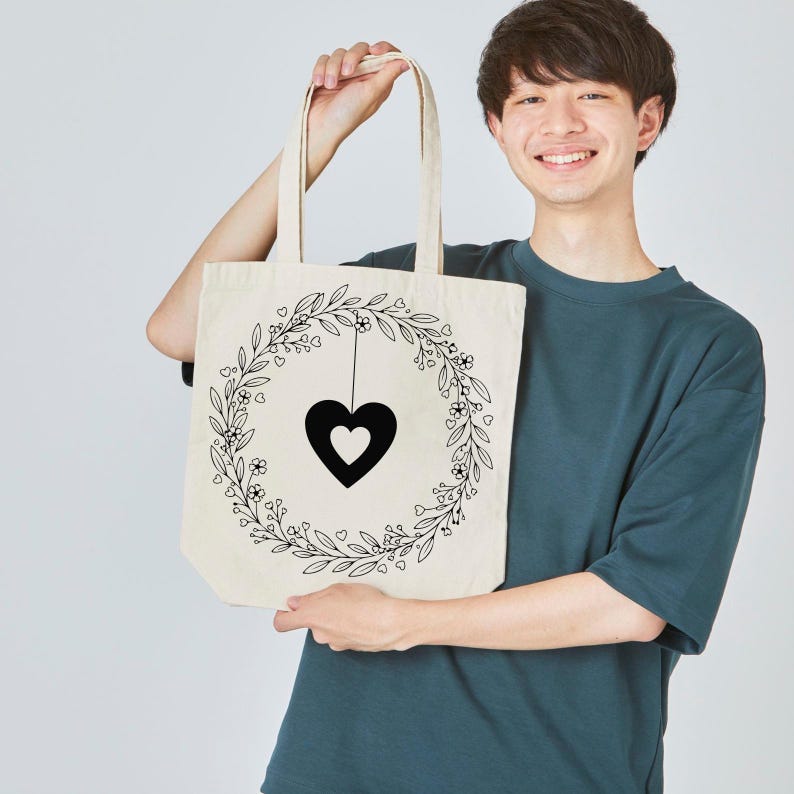 Peut inclure: Sac fourre-tout cr&egrave;me avec un pendentif en forme de c&oelig;ur noir &agrave; l'int&eacute;rieur d'une couronne florale. La couronne est compos&eacute;e de feuilles, de petites fleurs et de c&oelig;urs. Le sac a deux anses.